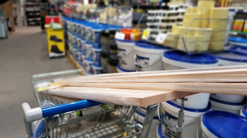 Personal perspective view of shopping cart with wooden boards and hardware supplies in store.